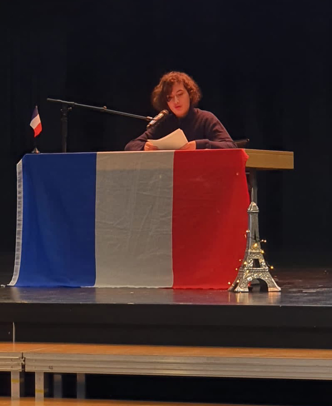 Schüler liest aus Buch an einem Tisch auf der Bühne, der mit französischer Flagge behangen ist, Eiffelturm-Modell steht daneben