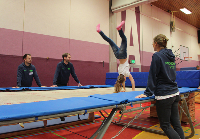 Mädchen turnt auf Trampolin, gesichert von Sportlehrern