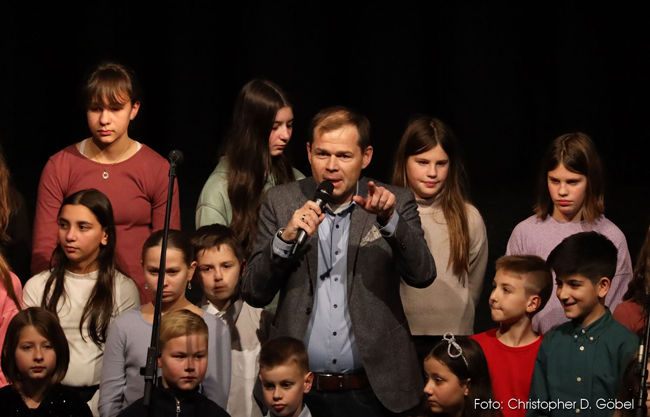 Stellvertretender Schulleiter mit Mikro zwischen Chorschülern auf der Bühne