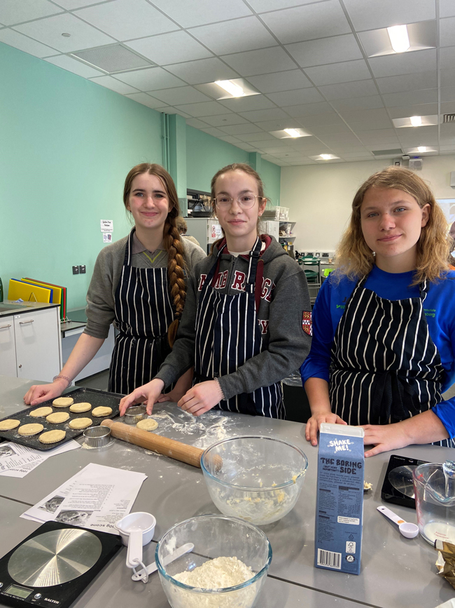 Schülerinnen beim Backen mit Backutensilien vor sich auf dem Tisch liegend lächeln in die Kamera, tragen schwarz-weiß gestreifte Schürzen