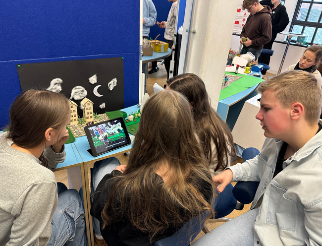 Schüler sitzen um ein Tablet, das eine Szene filmt, die sie davor aufgebaut haben