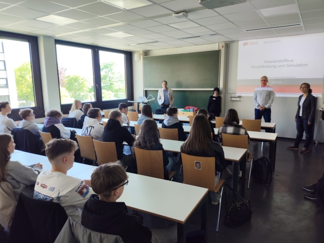 Schüler von hinten an langen Tischen sitzend, schauen auf Lehrer, die vor ihnen etwas erklären, Beamerbild im Hintergrund