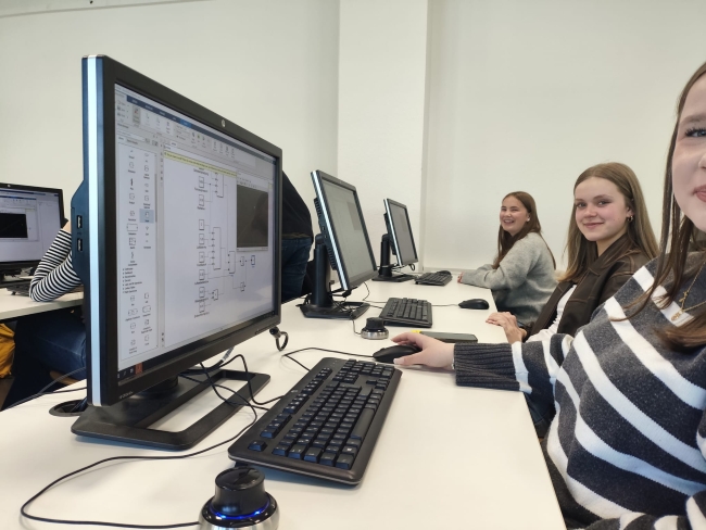 Drei Schülerinnen sitzen an PCs und schauen lächelnd in die Kamera