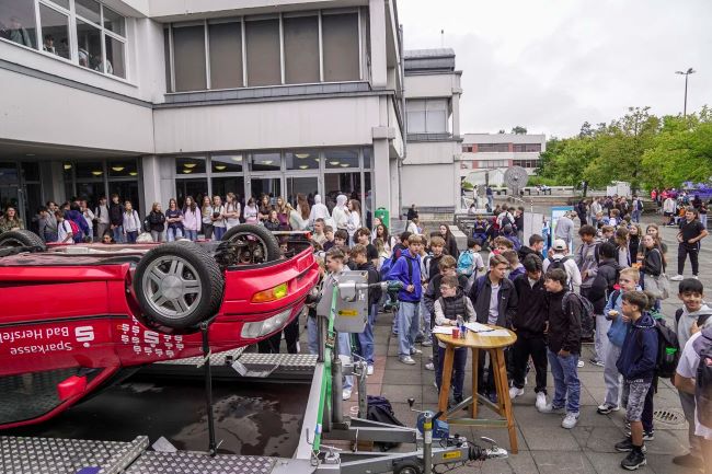 Schulhof mit vielen Schülern, im Vordergrund Auto, dass auf dem Dach liegt