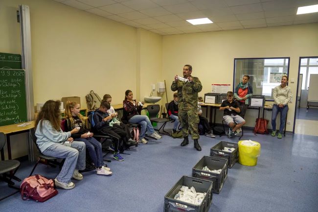 Klassenraum, in dem Schüler in einem Kreis sitzen; Bundeswehrsoldat erklärt etwas mit Verband in der Hand