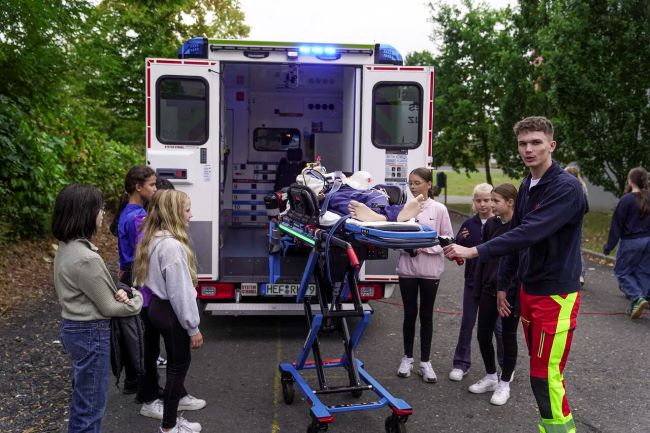Rettungssanitäter zeigt Schülern wie er gerade eine Patientenpuppe auf der Trage in den Wagen schiebt