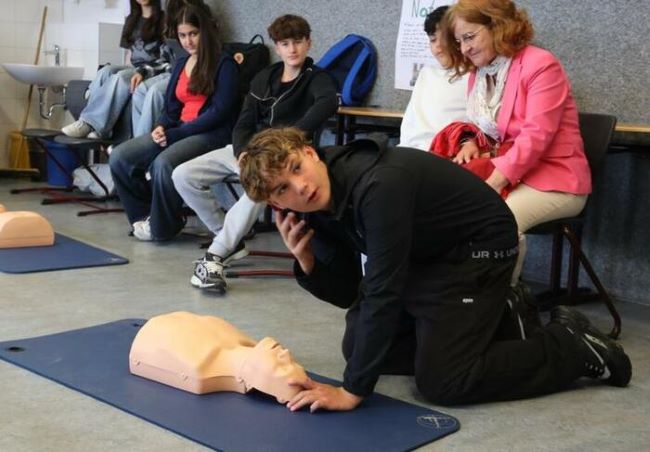Schüler kniet über einer Beatmungspuppe mit Telefon am Ohr, im Hintergrund sitzen Schüler und Erwachsene