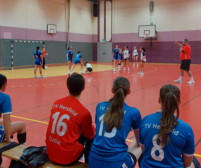 Drei Mädchen mit Trikots von hinten zu sehen sitzend schauen beim Spiel ihrer Mannschaft von der Seite aus zu