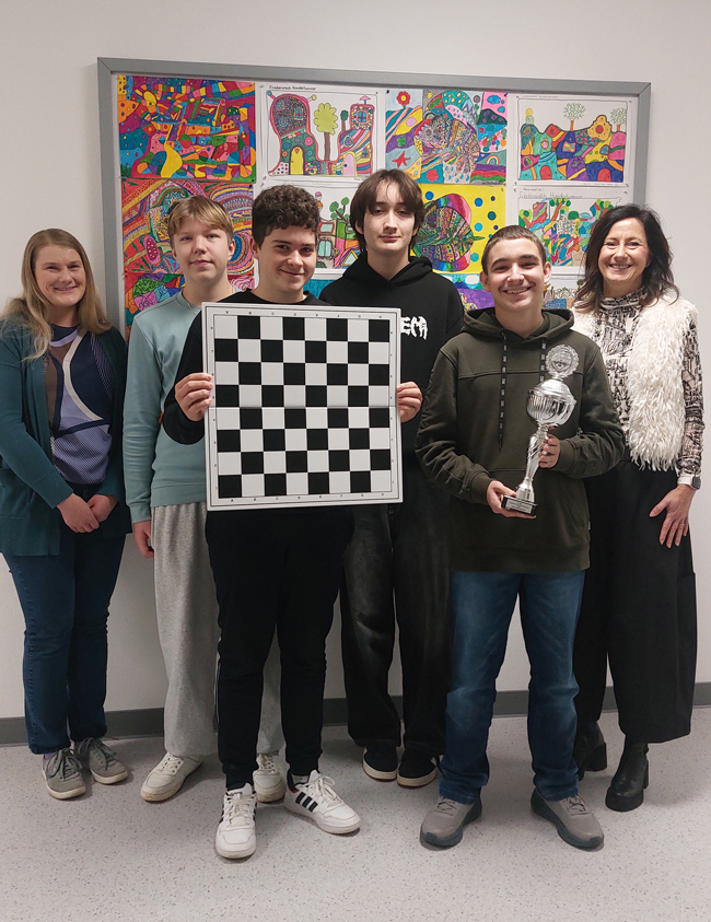 Vier Schüler mit Schachbrett und Pokal in der Hand umringt von zwei Frauen vor einem bunten Bild