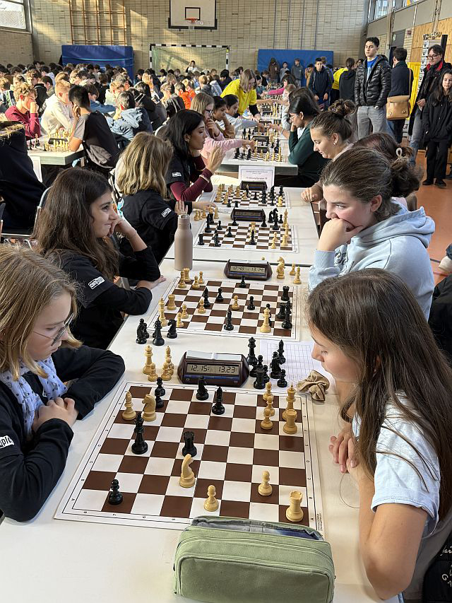 Mädchen an Schachbrettern in einer Turnhalle voller Tische mit Schachspielern