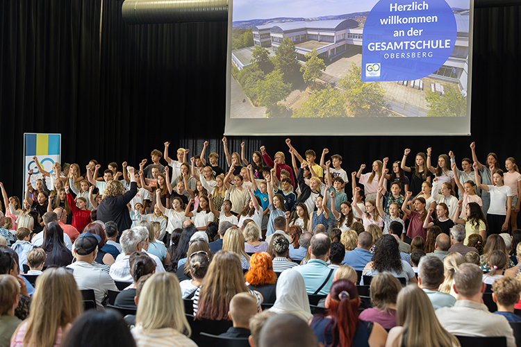 Chorschüler stehen zusammen auf der Bühne, singen und strecken dabei eine Hand in die Höhe; im Vordergrund Publikum und hinter dem Chor ist eine Leinwand mit einem Bild der Schule und der Aufschrift "Herzlich Willkommen an der Gesamtschule Obersberg"