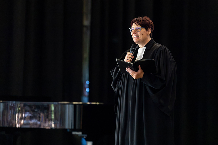 Schulseelsorgerin im Pastorinnengewand mit Mikro in einer und einem kleinen aufgeklappten Buch in der anderen Hand auf der Bühne zum Publikum sprechend