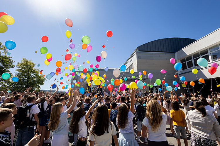 Luftballons steigen in blauen Himmel über dem Schulgebäude, Schüler und Eltern schauen ihnen begeistert und gespannt nach, viele filmen oder fotografieren mit Handys