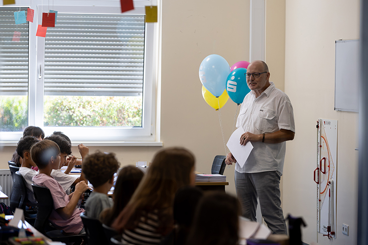 Blick in Klassenraum, in dem Luftballons an der Decke hängen; Lehrer steht vor den Schülern neben der Tafel und erklärt etwas
