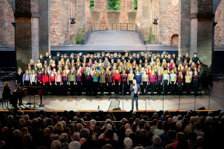Blick vom Publikum aus auf große Bühne herunter, die voller Sänger (in vier Reihen stehend) steht, mit Klavier links und vor ihnen redet ein Mann zum Publikum