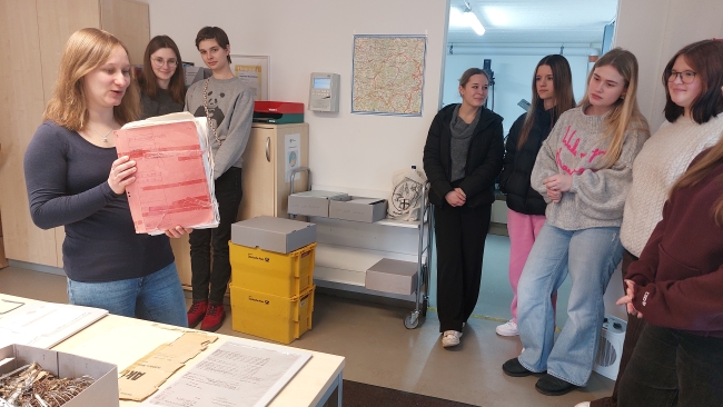 Frau erklärt Schülerinnen, die um sie herum stehen, etwas mit einer alten Mappe in der Hand