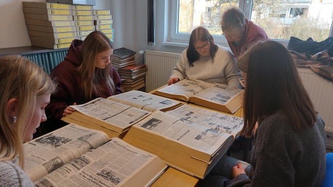 Vier Schülerinnen sitzen an einem Tisch mit A3-großen Büchern voller alter Zeitungen, eine Lehrerin schaut ihnen über die Schultern