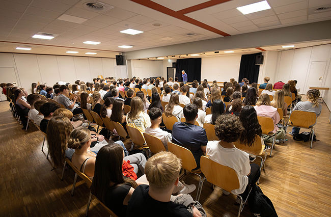 Saal mit Publikum auf Stühlen sitzend von hinten mit Blick auf einen kleinen Mann im Hintergrund, der zum Publikum spricht