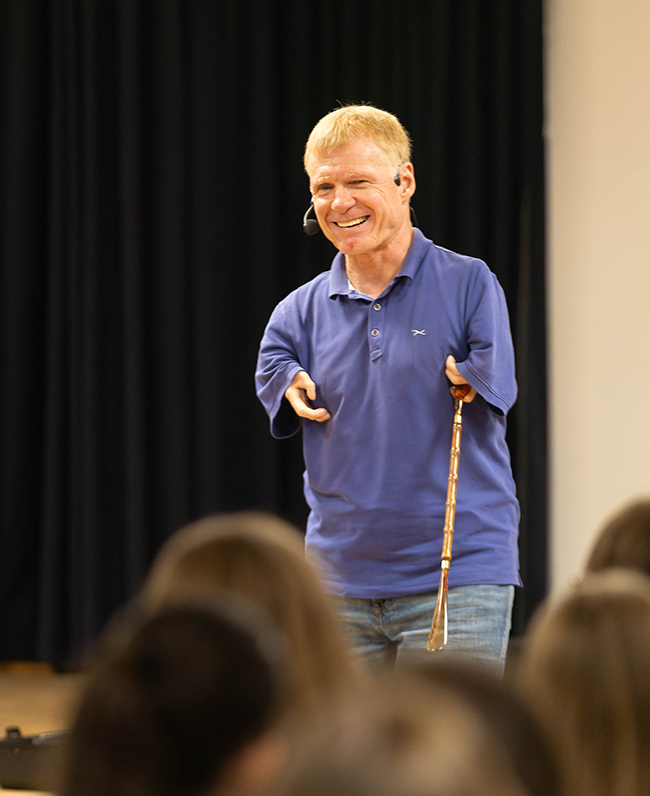 Der Redner lächelnd mit Kopfmikrofon und einem Stock in der linken Hand, Hände beginnen unmittelbar an den Schultern und schauen gerade so aus den Hemdärmeln seines blauen Poloshirts heraus
