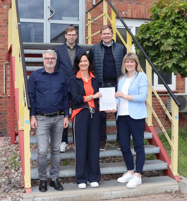 Fünf Personen auf einer Treppe vor einem Eingang zu einem Backsteinhaus; zwei Frauen mit Urkunde in der Hand