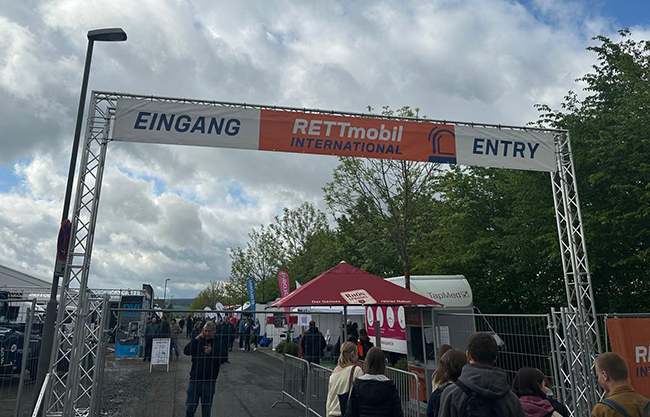 Eingangstor zur Messe durch das Schüler hindurchgehen, im Hintergrund Messestände und Zelte, bewölkter Himmel