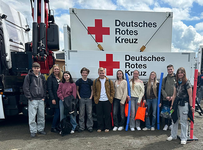 Gruppenfoto vor einem großen Plakat mit der Aufschrift Deutsches Rotes Kreuz, das an der Seitenwand eines Trucks hängt, Schüler teils mit Beuteln und Schwimmnudeln