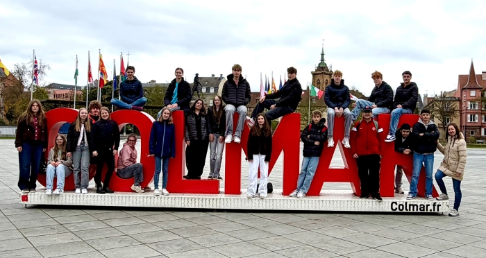 Schüler und Lehrerinnen stehen und sitzen verteilt auf und um lebensgroßen, roten Buchstaben, die den Begriff Colmar bilden
