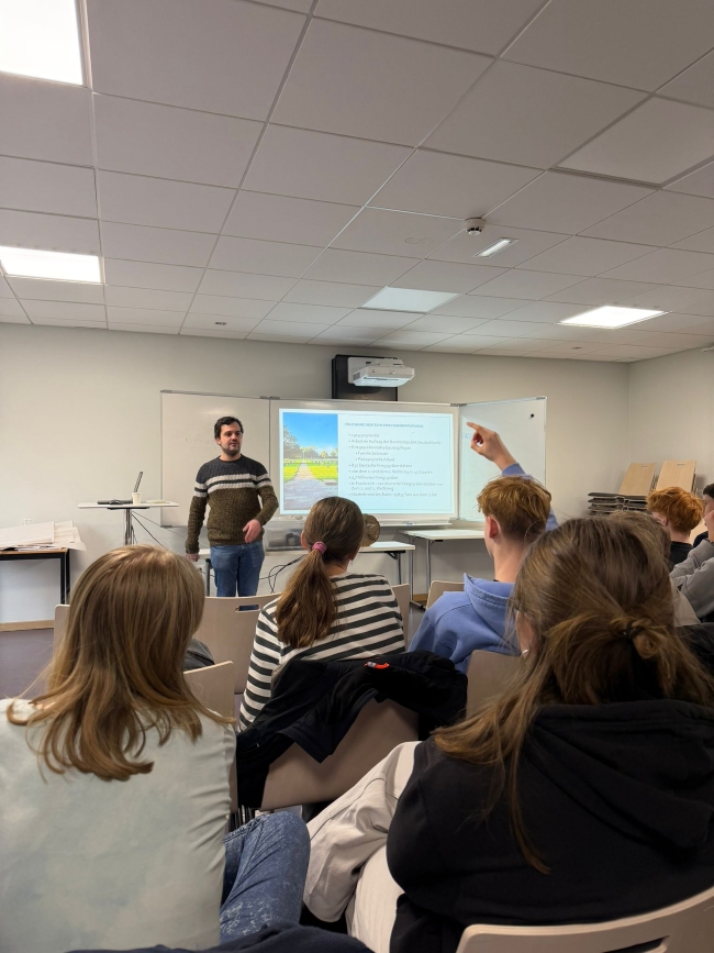 Schüler in einem Raum sitzend, schauen auf ein Beamerbild an der Wand. Lehrer vorne reagiert auf einen Schüler, der die Hand hebt