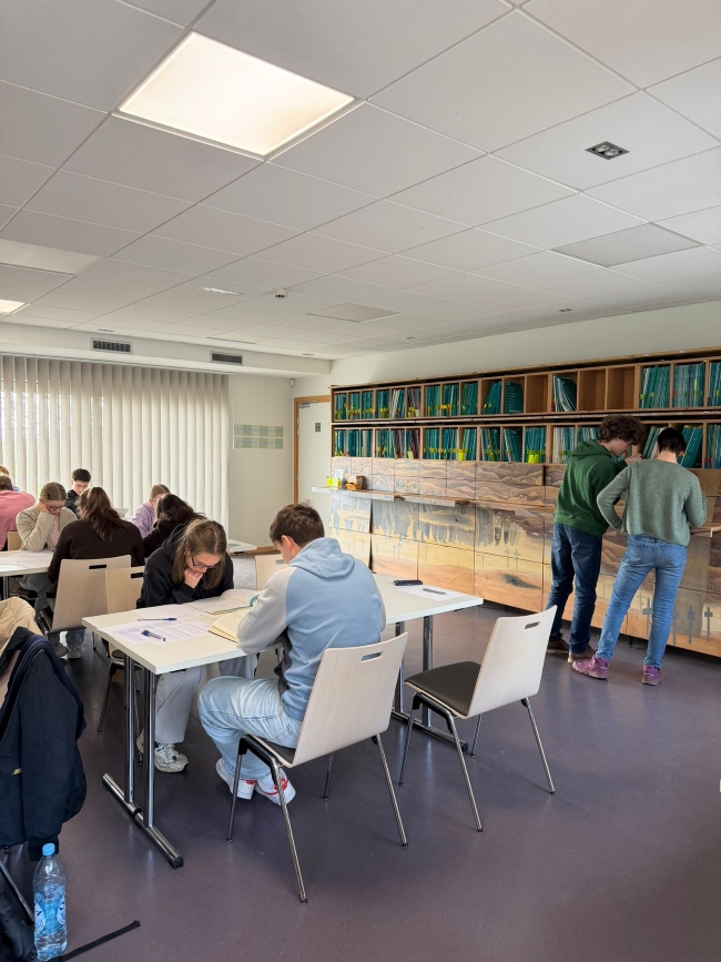 Schüler an Tischen in einem Raum sitzend recherchieren in Büchern oder suchen sich Bücher aus einem Regal