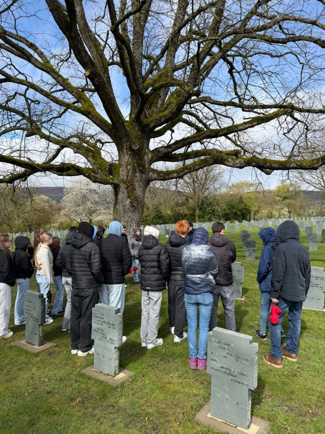 Schüler von hinten mit gesenkten Köpfen andächtig auf einem Friedhof stehend mit einem großen kahlen Baum, der über ihre Köpfe neigt