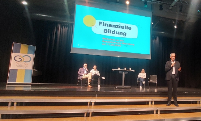 Bühne auf der ein Banner mit GSO-Aufschrift, ein Tisch mit drei Personen, die drum herum sitzen und eine Leinwand zu sehen ist, auf der der Titel der Veranstaltung "Finanzielle Bildung" steht. Davor steht ein Mann und spricht zum Publikum, das nicht zu sehen ist