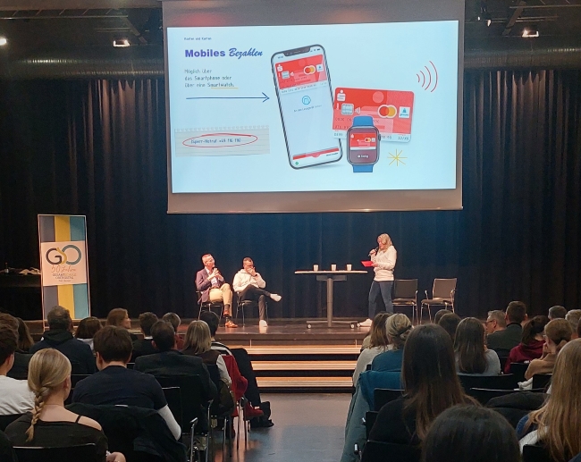 Wieder Blick aus Publikum auf Bühne, wo Frau spricht; auf der Leinwand sind ein Handy, eine Smartwatch und eine Geldkarte der Sparkassse zu sehen