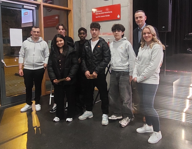 Sparkassen-Mitarbeiter und Schüler stehen zusammen vor der Bühne mit Blick in die Kamera