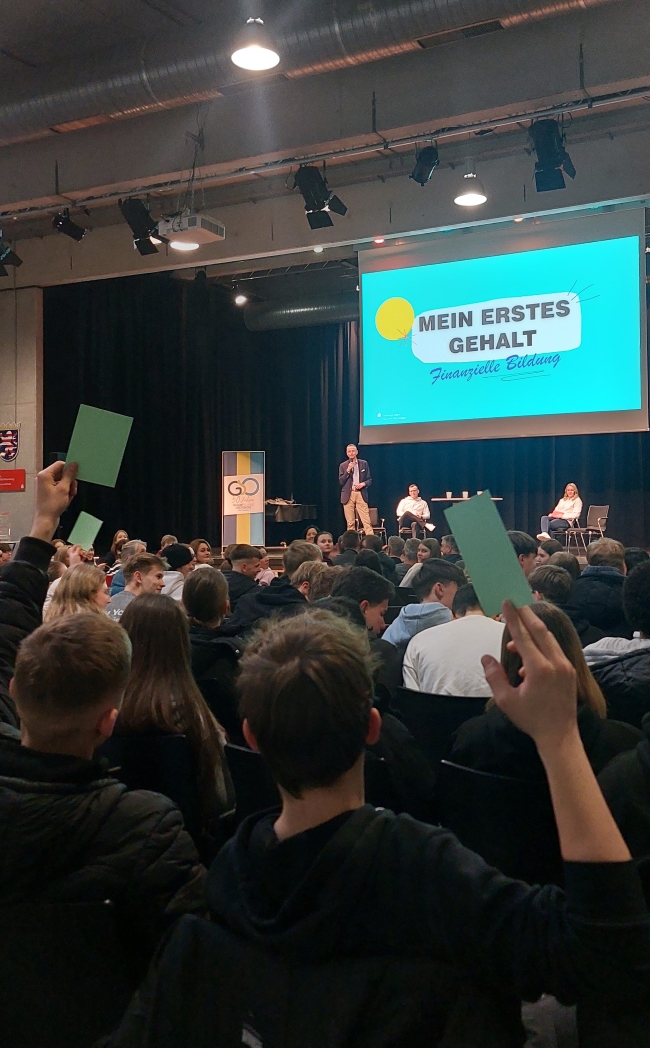 Schüler heben Karten nach oben, sind von hinten mit Blick auf die Bühne zu sehen