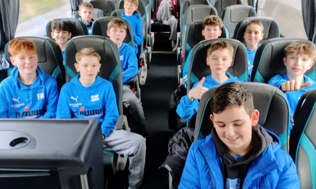 Schüler im Bus in Zweierreihen sitzend mit hellblauen Pullovern, lächeln in die Kamera und halten teils Daumen hoch