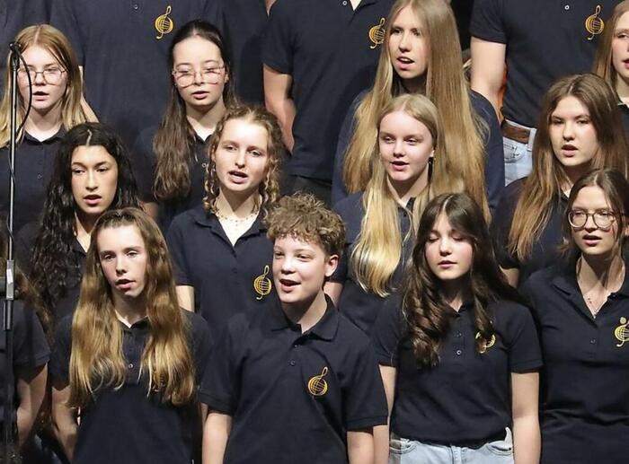 Nahaufnahme von Schülern, schwarz gekleidet in drei Reihen gestuft hinter einander stehend auf der Bühne, die singen