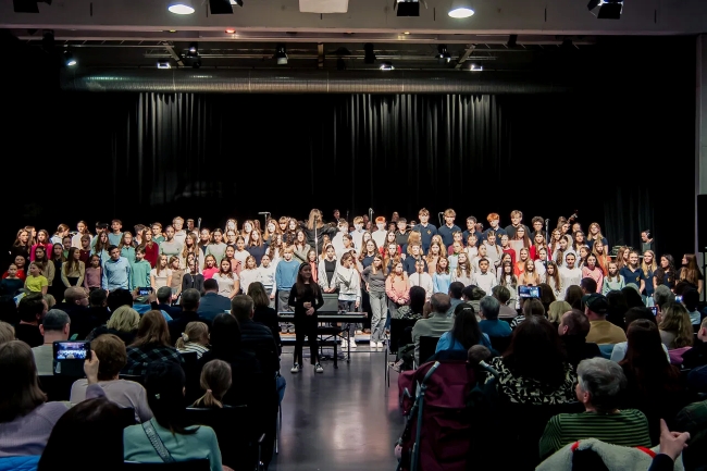 Blick von weiter hinten im Saal auf Publikum von hinten und großen Chor mit bunt gekleideten Schülern auf der Bühne singend