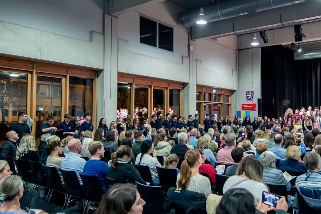 Blick in den Saal mit Schülern die links neben Publikumsreihen stehen und singen