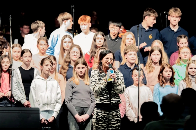 Schulleiterin spricht in Mikro, lächelnd vor Chorschülern stehend, zum Publikum