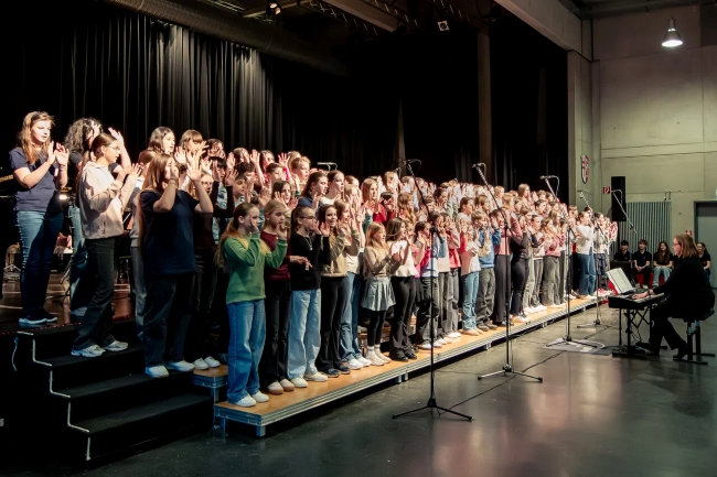 Chor mit zahlreichen Schülern auf der Bühne von links neben der Bühne betrachtet, gestikulierend singend