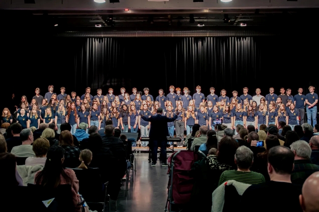 Chor von weitem fotografiert mit Chorleiter von hinten im Mittelpunkt, Reihen des Publikums sind zu sehen