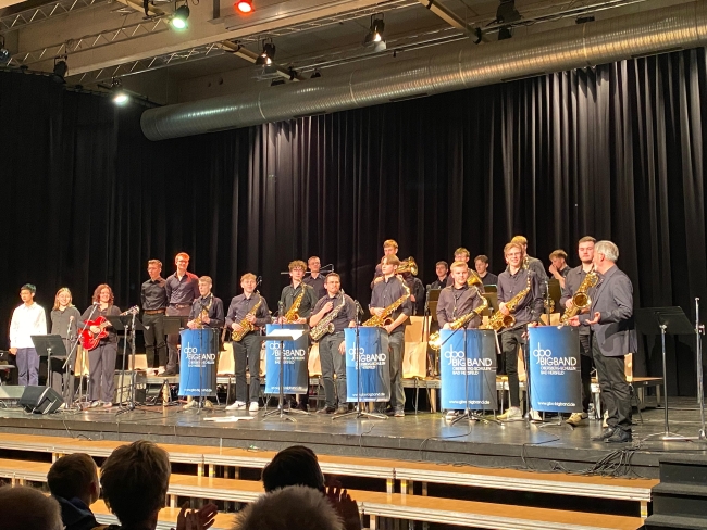 Orchester stehend hinter Notenständern mit blauen Bannern, auf denen groß GBO steht