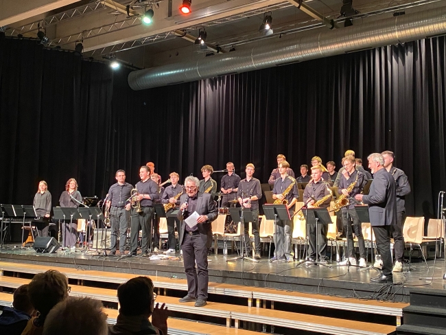 Orchester stehend auf der Bühne zum Publikum schauend, davor steht ein Mann moderierend