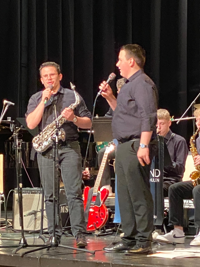zwei Schüler, einer mit Saxophon in einer Hand, halten Mikros und singen auf der Bühne vor dem Orchester