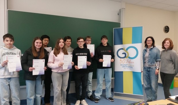 Schüler und zwei Lehrerinnen in einem Klassenraum vor einer Tafel und einem Banner mit Schullogo stehend, haben Urkunden in der Hand und lächeln