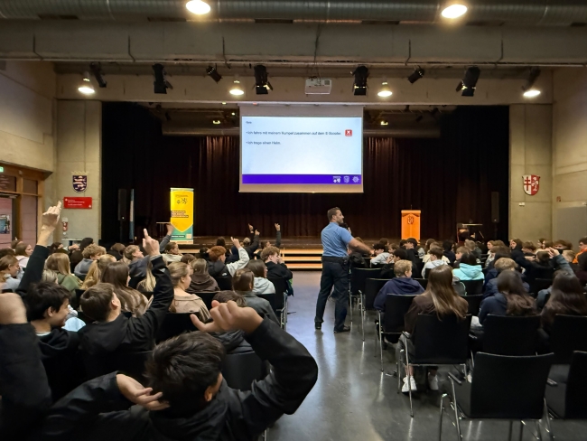großer Vortragsraum mit Blick auf Bühne mit großer Leinwand, auf der etwas steht; im Publikum im Vordergrund läuft ein Polizist mit Mikro