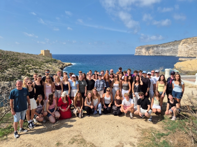 Schülergruppe vor malerischer Kulisse an einer Küste einer Insel mit Blick auf das blaue Meer, blauen Himmel und eine Steilküste auf der einen und gegenüberliegend eine Festung auf der anderen Seite