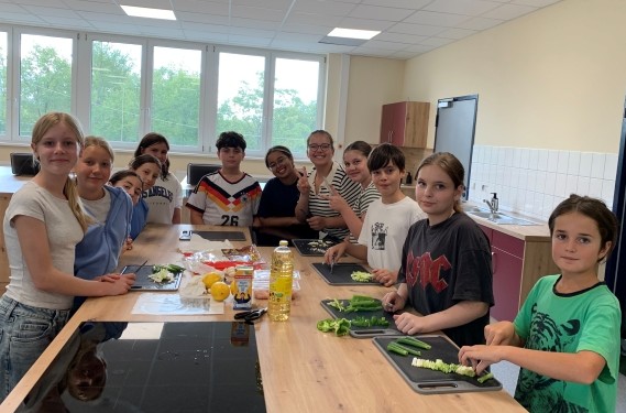 Schüler stehen um einen großen Küchentisch herum, schauen zur Kamera und sind gerade dabei Gemüse zu schneiden
