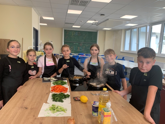 andere Schülergruppe an einem großen Küchentisch, teils mit Schürzen bekleidet; in der Tischmitte liegt frisch geschnittenes Gemüse, an der Tafel im Hintergrund stehen Rezepte