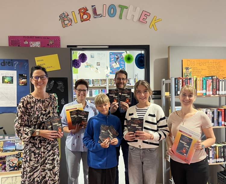 Erwachsene und zwei Kinder halten Bücher in die Kamera vor der Bibliothek stehend mit vielen Bücherschränken im Hintergrund